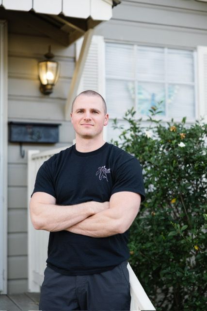 Man in black t-shirt with logo, smiles in front of a house, standing on porch with hand on railing.