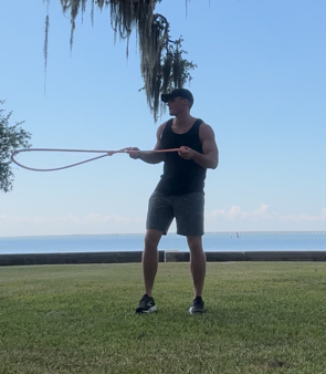 Man exercising with a resistance band outdoors. He is standing on grass, by the water, holding the band.