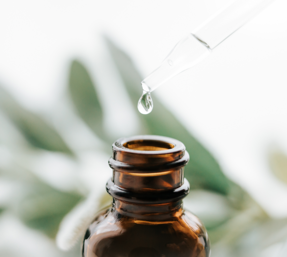 Brown dropper bottle with a drop of liquid falling, background blurred green leaves.