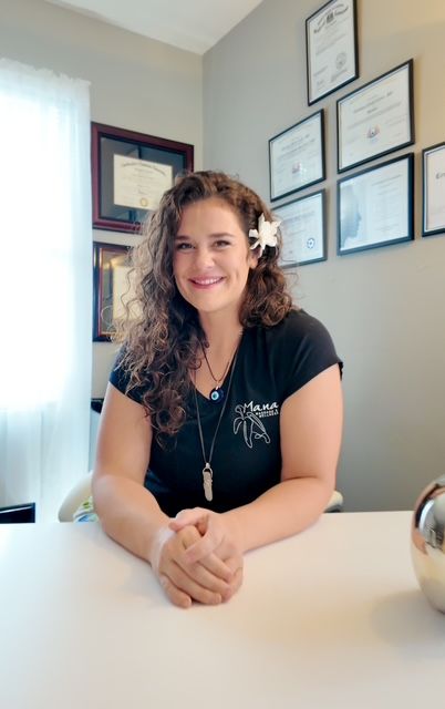 Woman with curly brown hair, flower, black shirt, smiling, sitting at desk in office with diplomas.