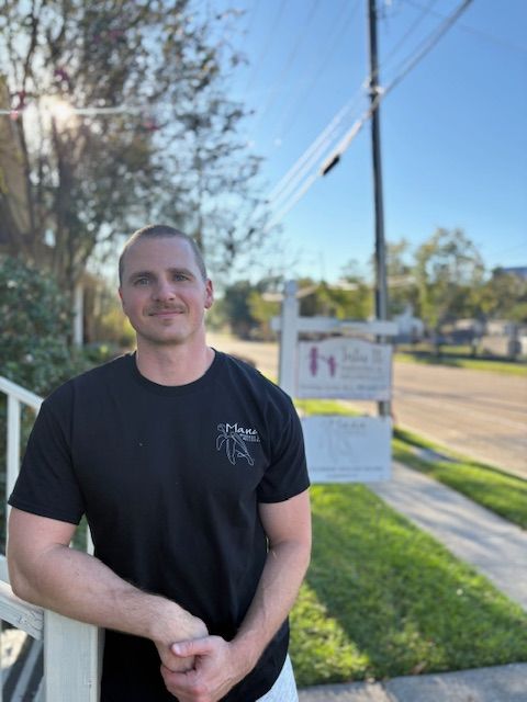 Man in black t-shirt smiles outside a building with a sign. Sunny day, leans on a railing.