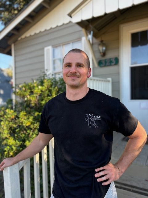 Man in black t-shirt with logo, smiles in front of a house, standing on porch with hand on railing.