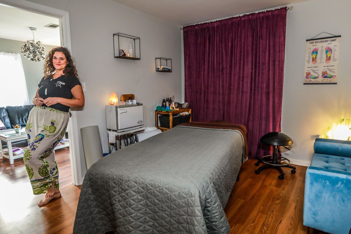 Woman in massage room leans against doorway. Massage table, blue chair, purple curtain.
