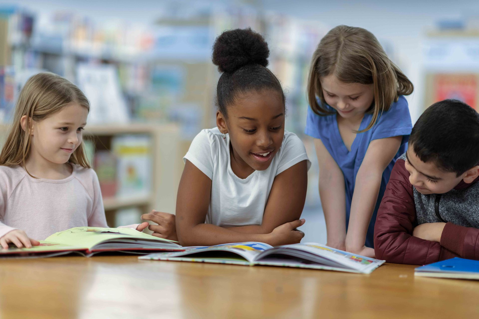 Four children look down at open books on a wooden table in a library setting.