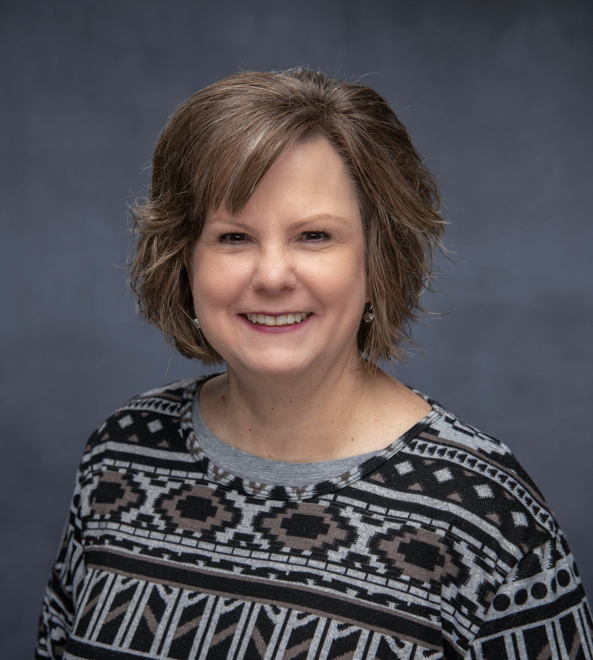 A smiling woman with shoulder-length brown hair, wearing a black-and-white patterned top against a dark blue background.