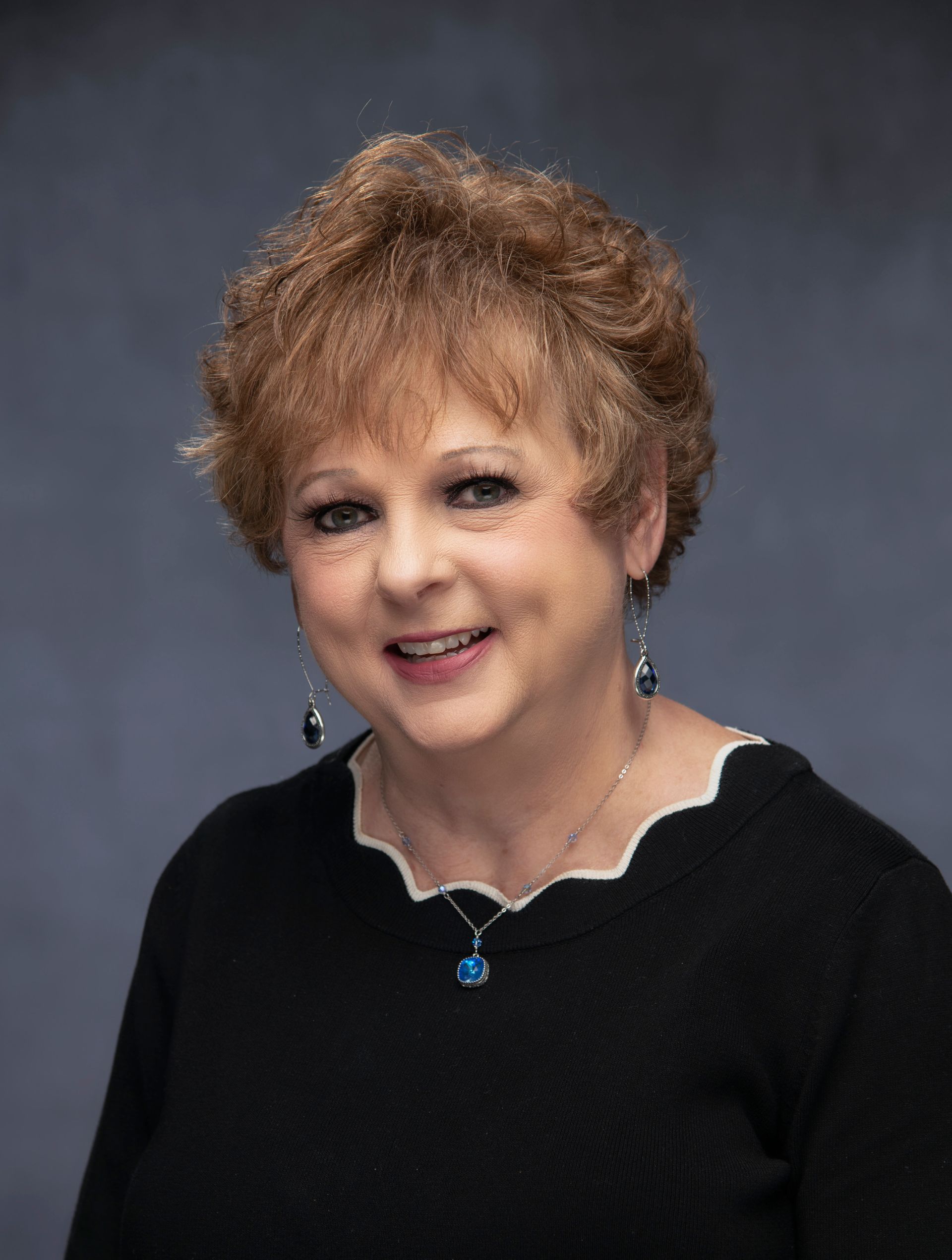 A headshot of a smiling person with curly, light brown hair, wearing a black shirt with a scalloped neckline and earrings.