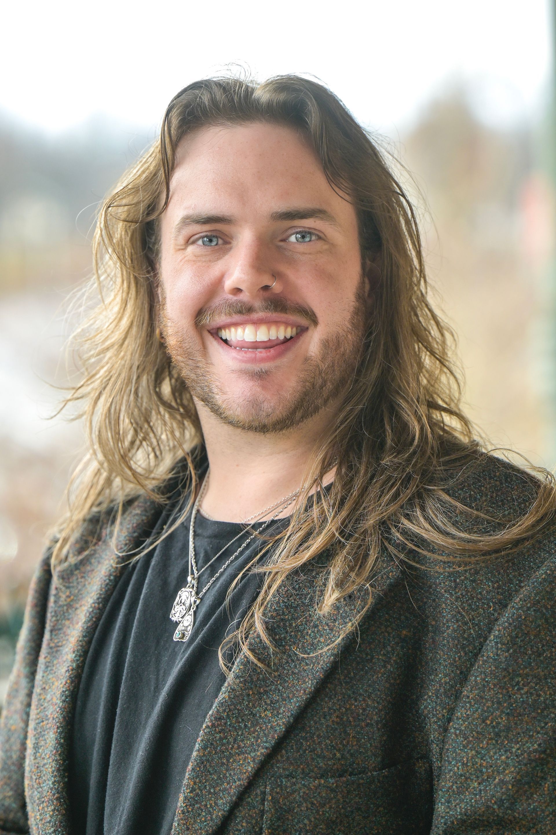 Smiling person with long, wavy brown hair, wearing a dark textured blazer and a silver pendant necklace.