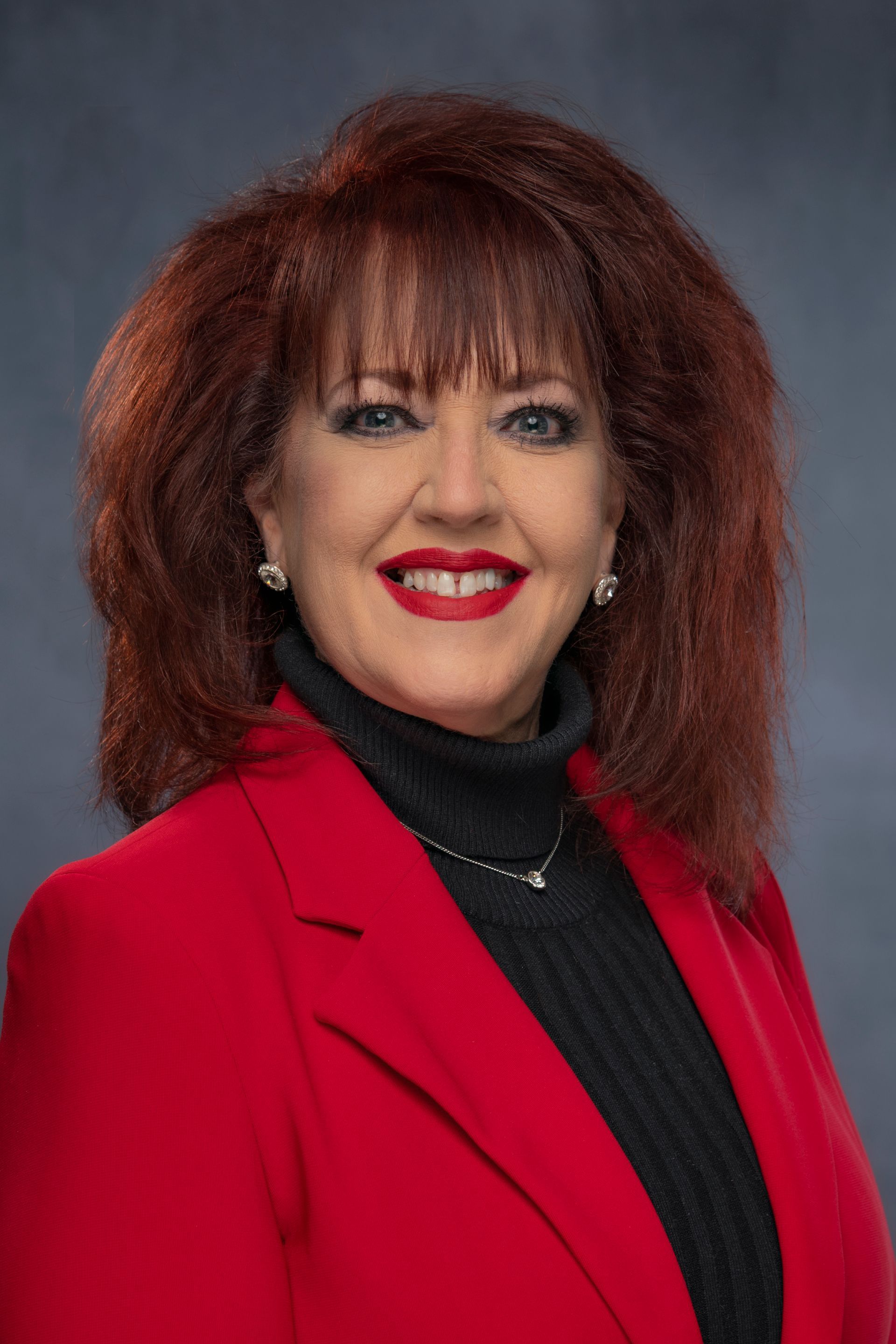 A professional portrait of a person with auburn hair wearing a red blazer and a black turtleneck against a gray background.