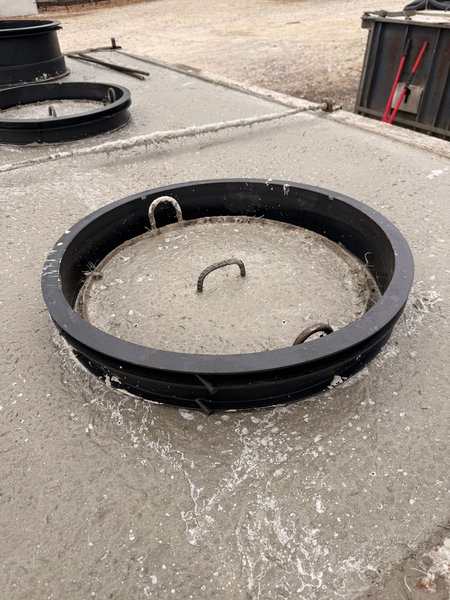 Circular metal manhole cover with lifting handles sits on concrete.