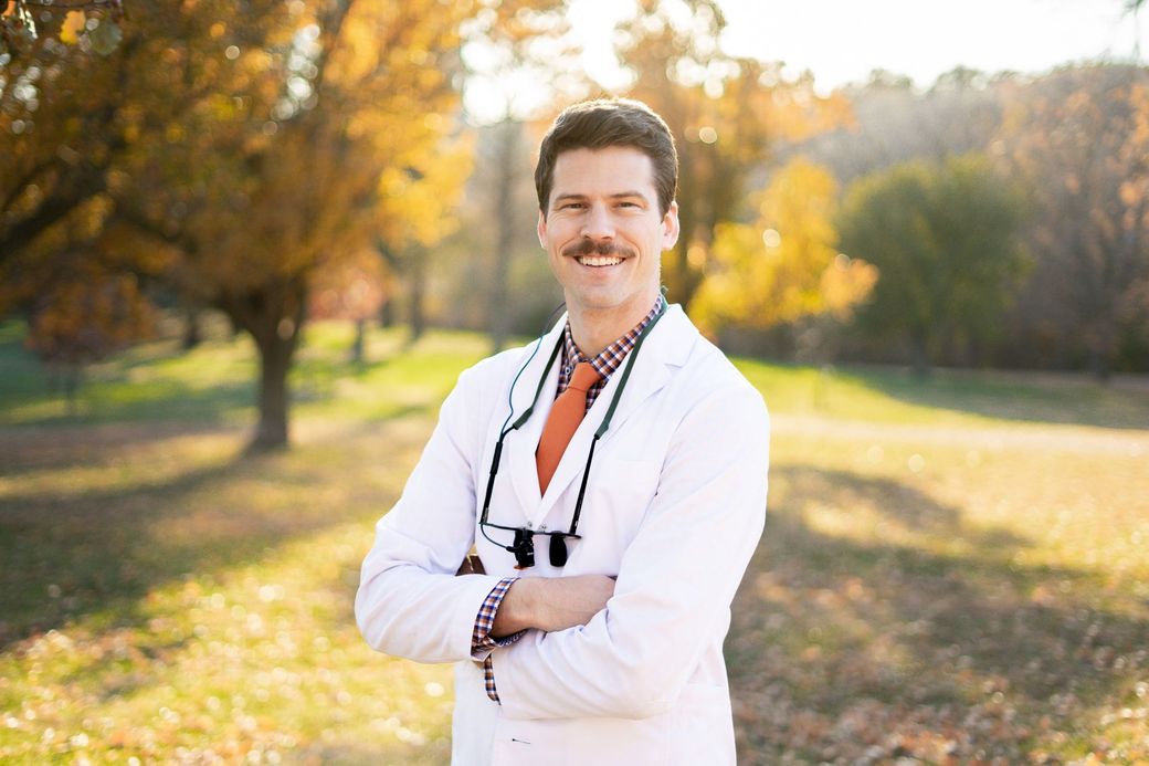 Man in lab coat with arms crossed, standing outdoors in autumn.