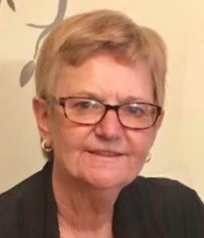 A person with short, light hair and glasses, wearing a black top, smiling against a light-colored wall with floral decor.