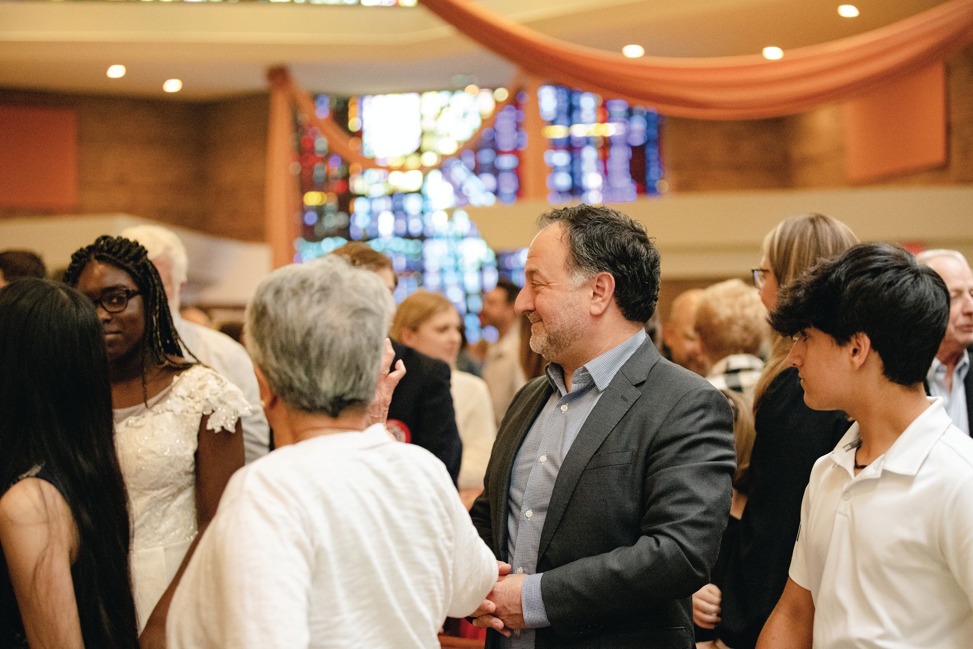 People greet each other at an indoor social gathering with warm lighting and decorative drapes in the background.