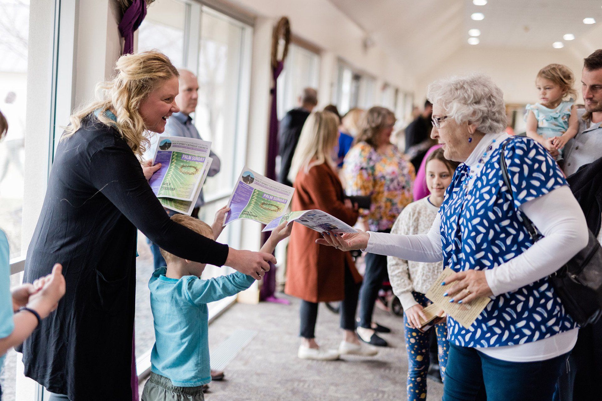 A person in a black sweater hands paper programs to a child and an adult in a patterned blue shirt in a bright hallway.