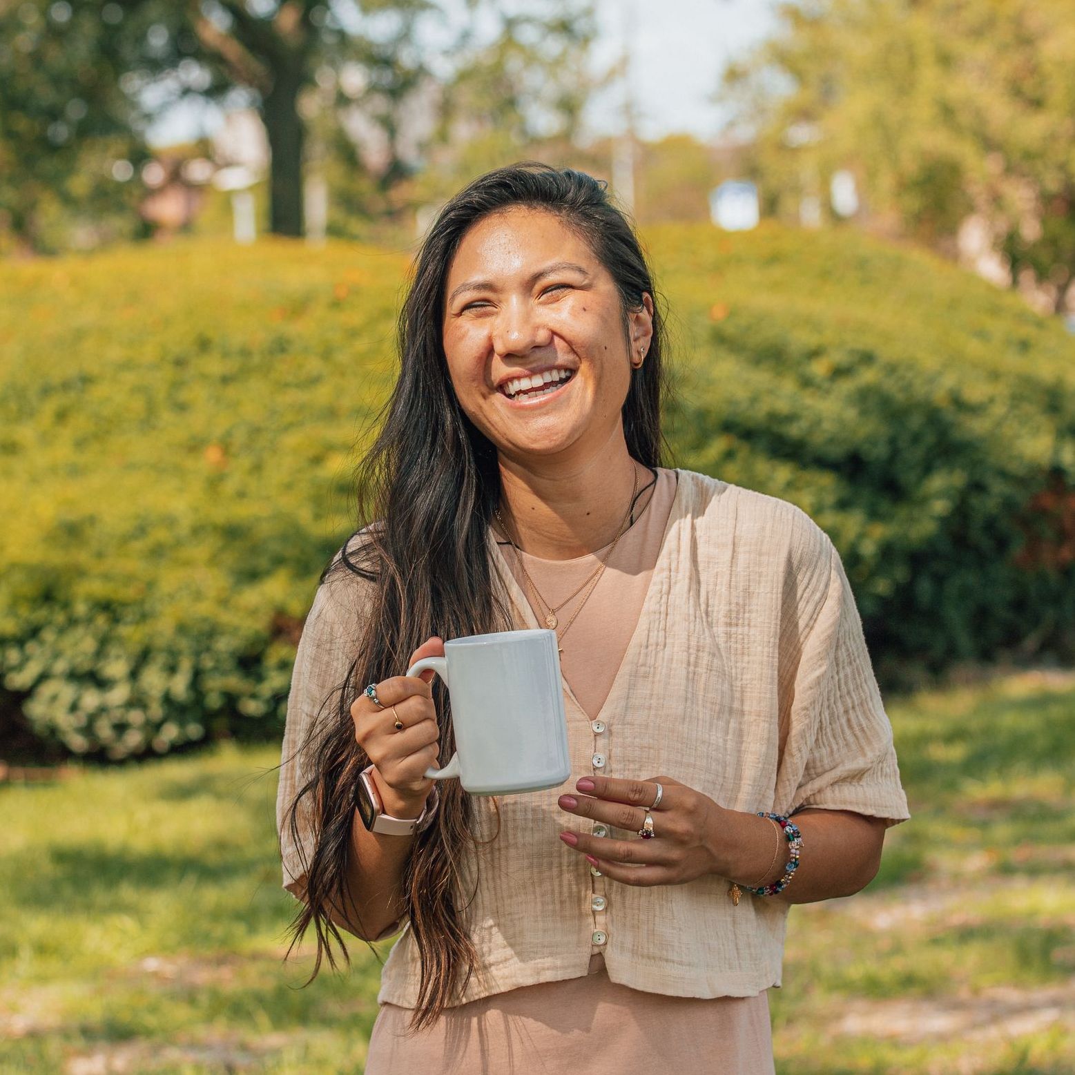 A smiling person with long dark hair holds a white mug while wearing a tan shirt, standing in a sunny, grassy park.