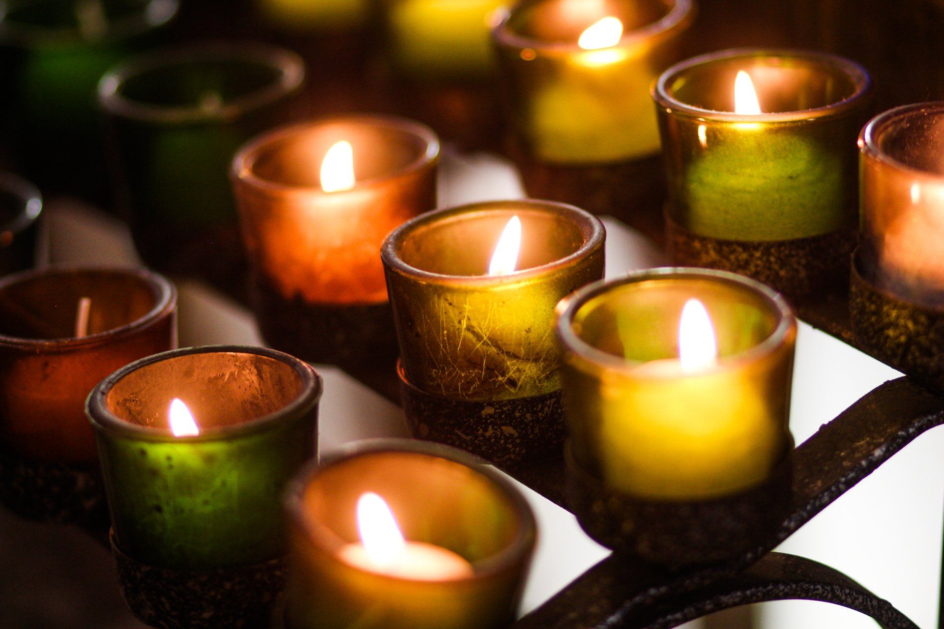 Multiple glowing votive candles in colored glass holders sitting on a metal rack.