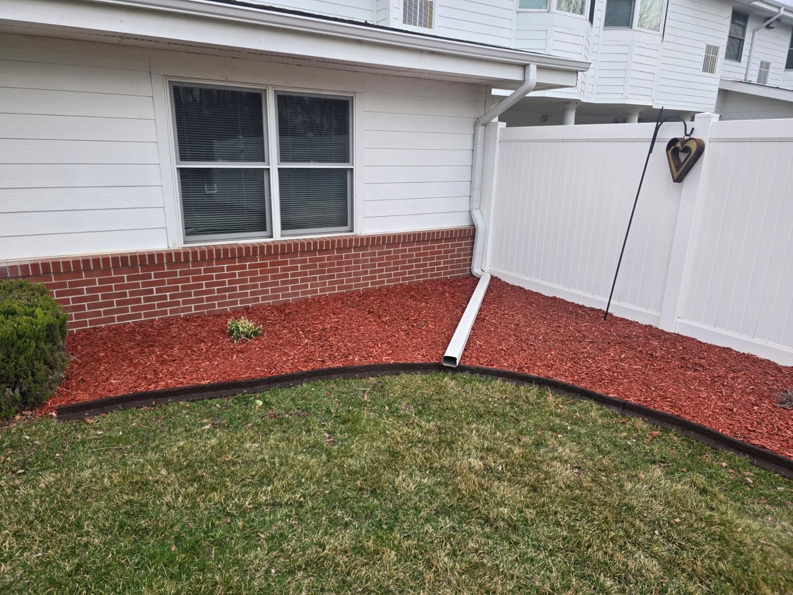 A white house corner with red mulch, a brick base, a downspout, and a white privacy fence bordering a lawn.