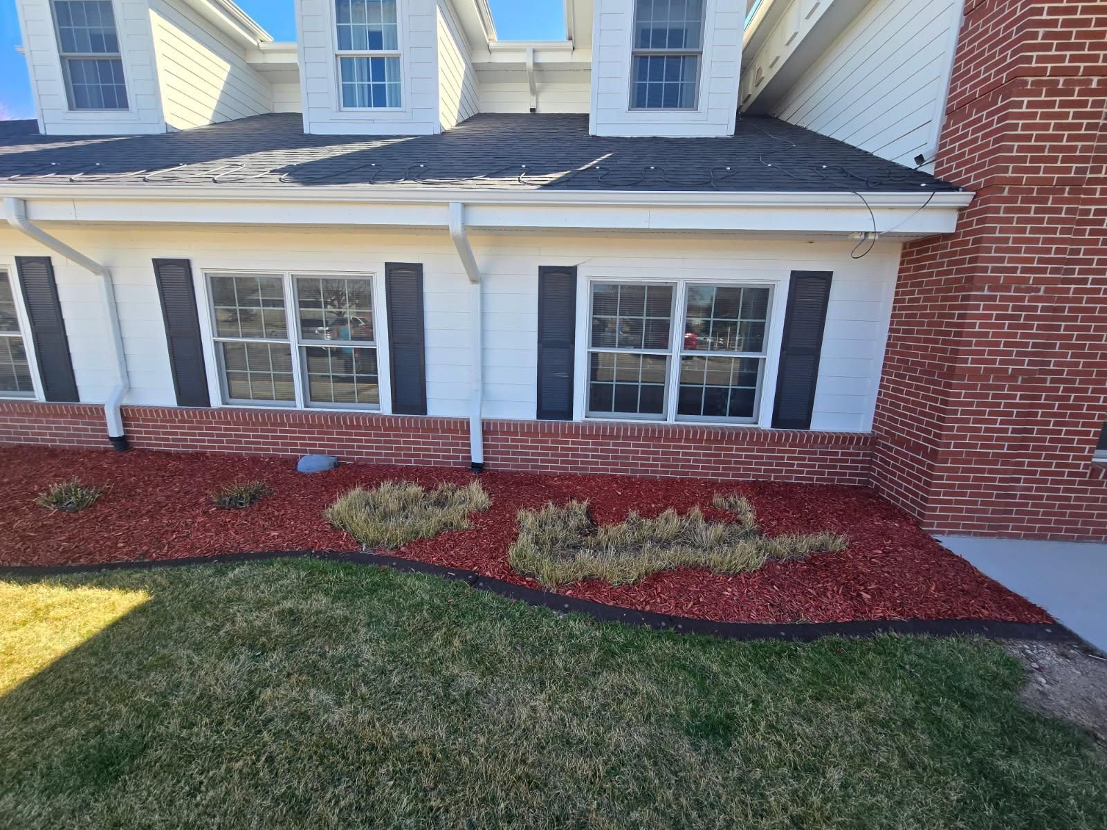 A white house exterior featuring two windows with black shutters, a brick base, and red mulch landscaping under a lawn.