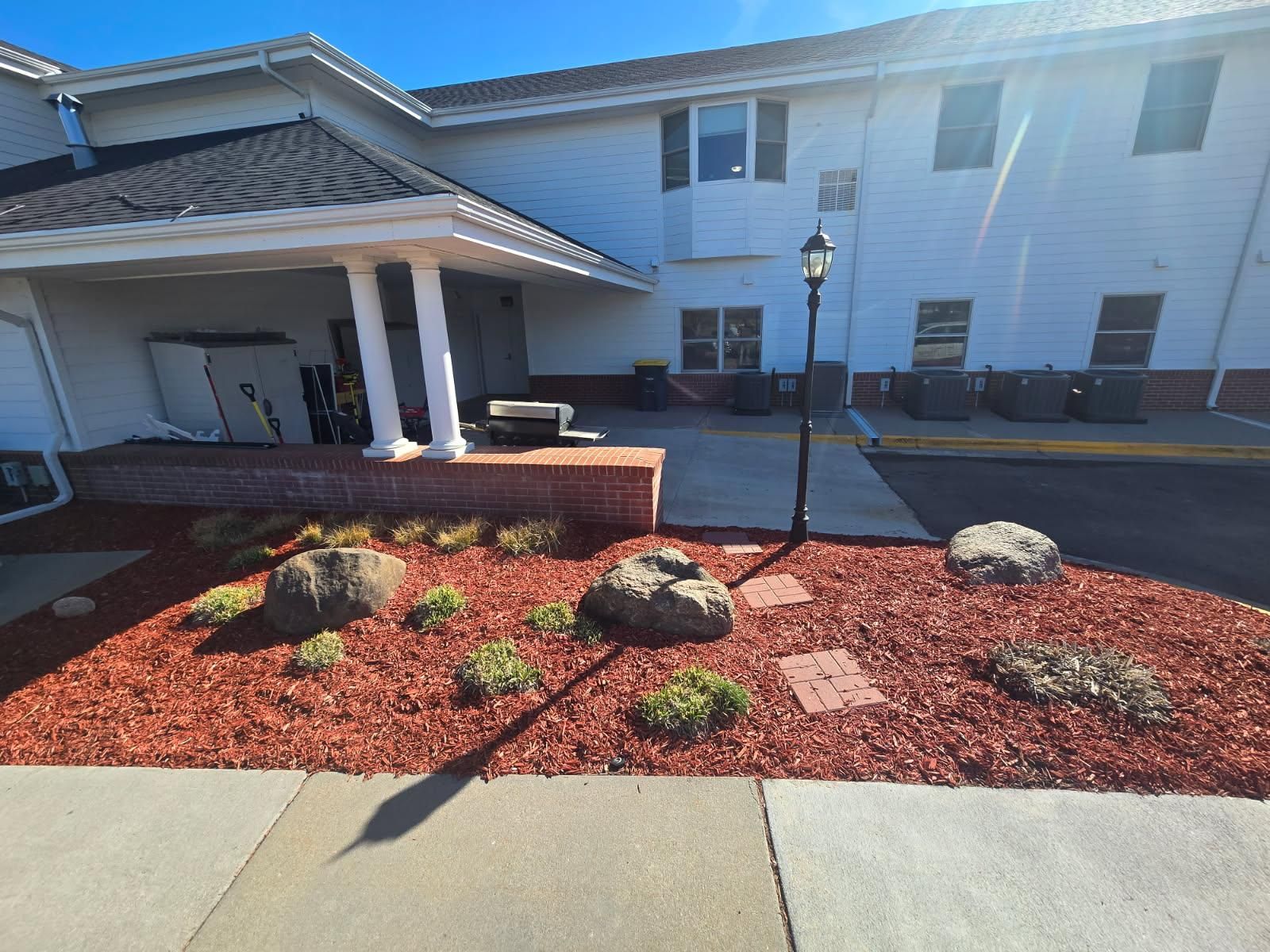 A light-colored building with a covered entrance and a front garden bed featuring large rocks, small plants, and red mulch.