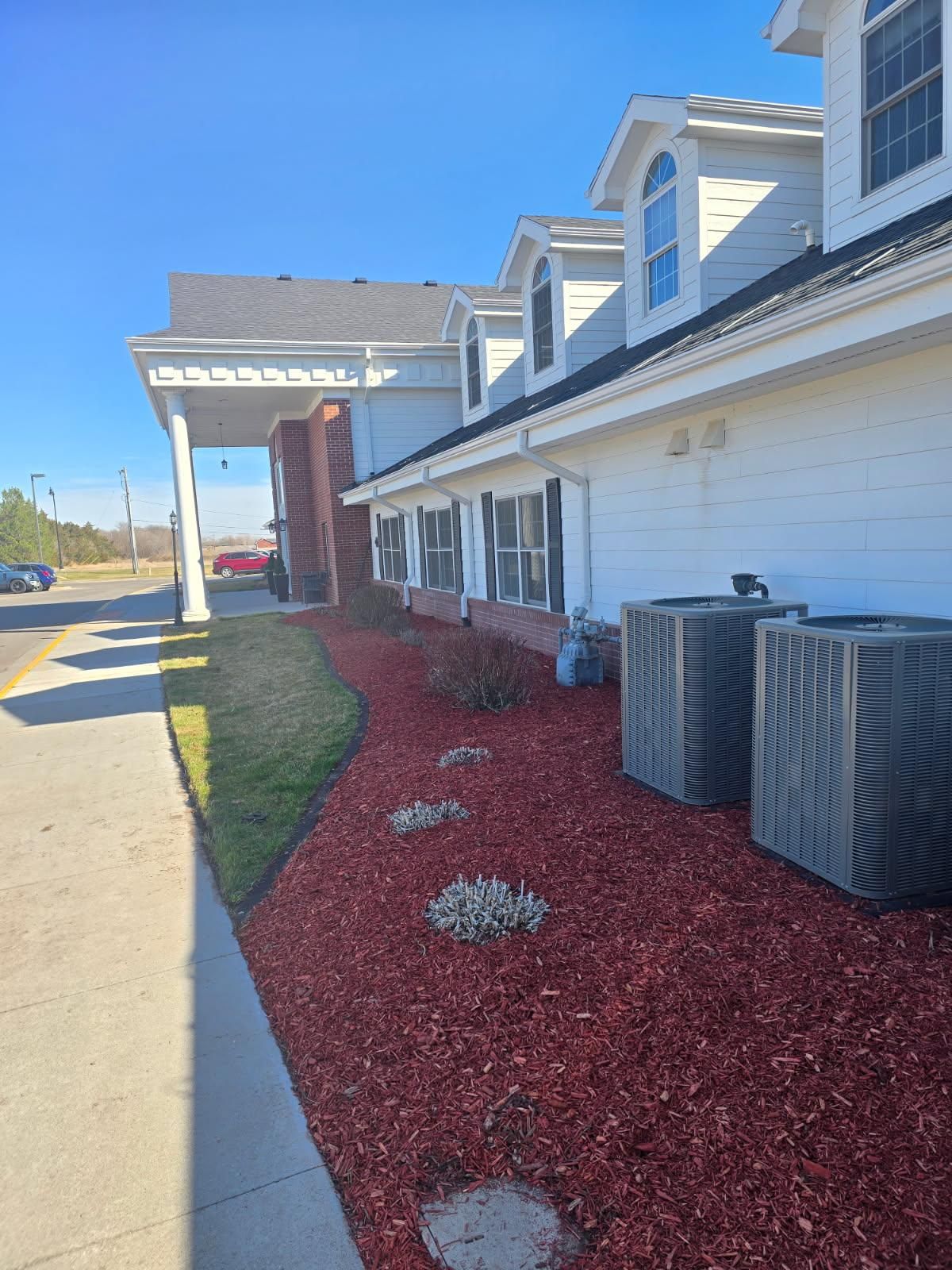 A white building with a gray roof and red mulch landscaping features, seen from the sidewalk on a sunny day.