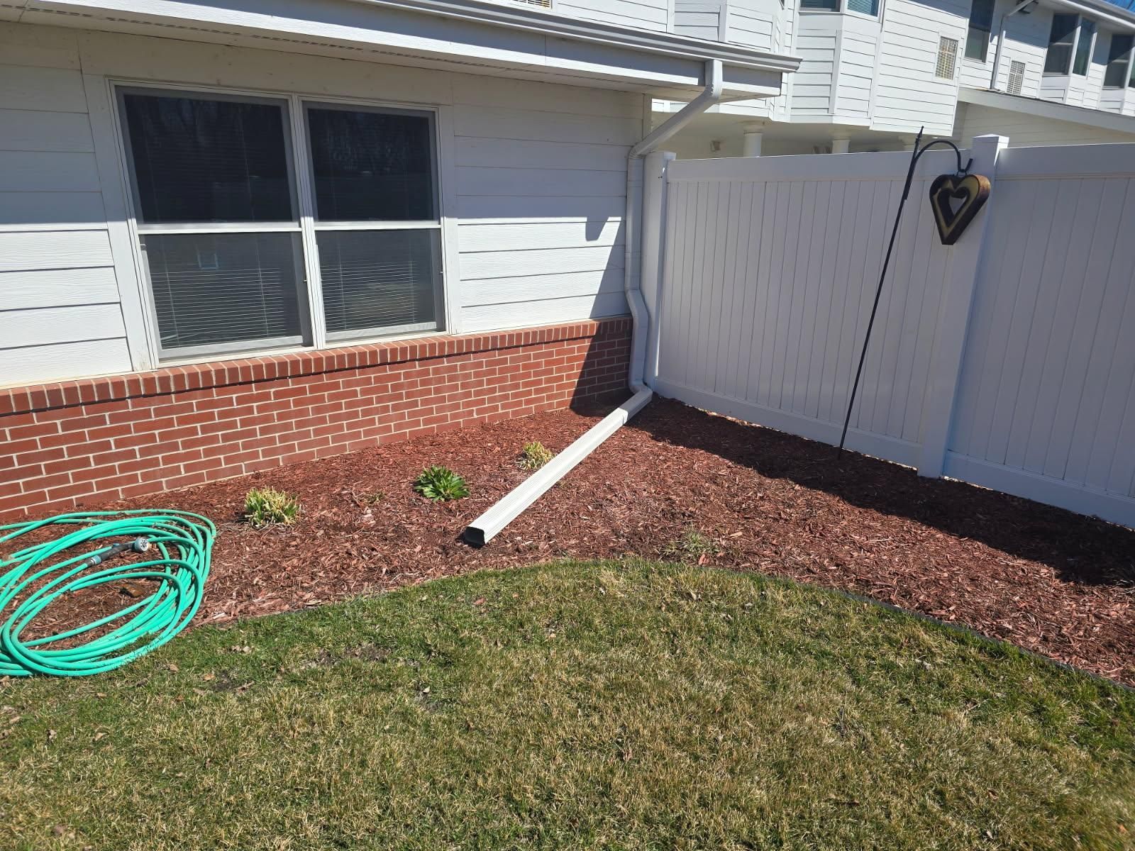 A white gutter downspout extends from a house into a landscaped yard with red mulch, brick skirting, and a white fence.