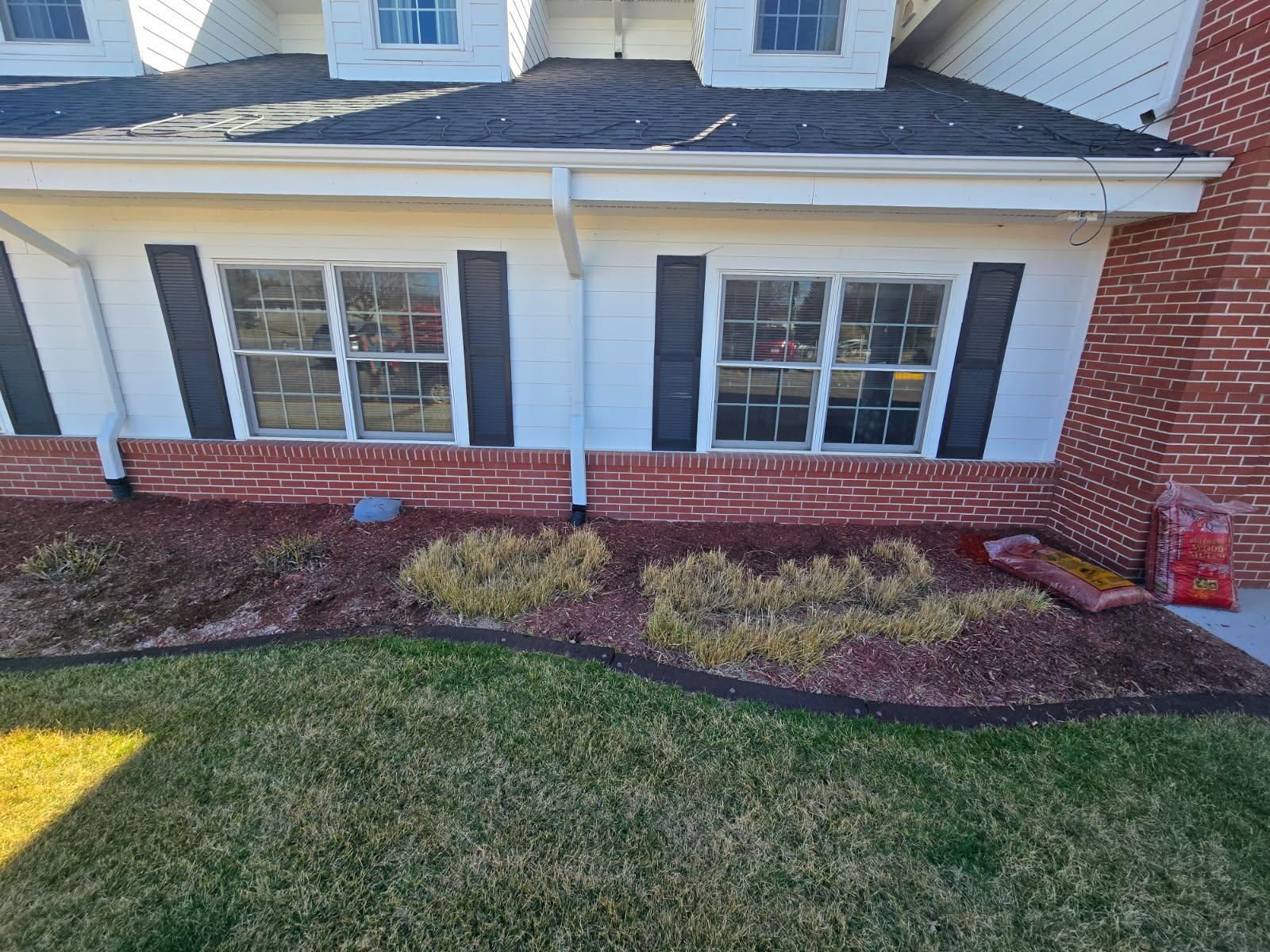 A brick and white home exterior with two windows, dark shutters, and a mulched garden bed in the front yard.