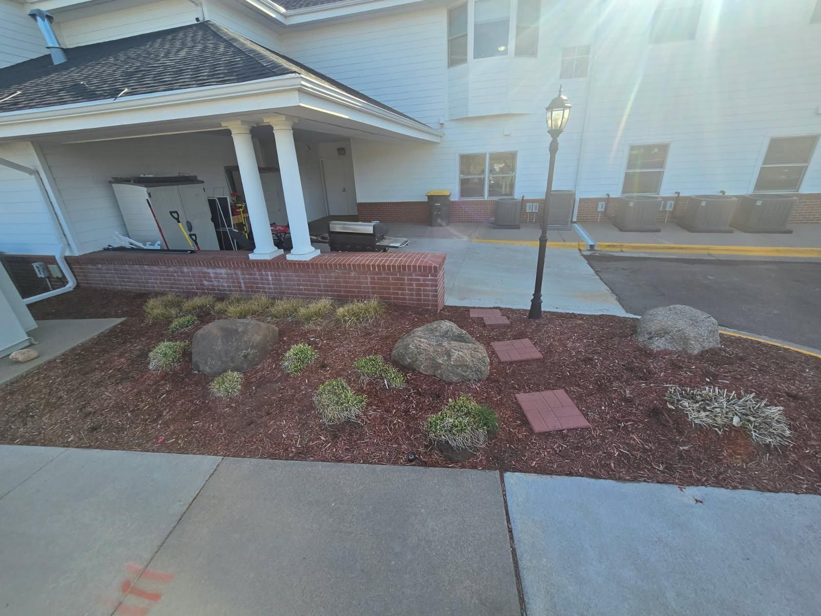 A garden bed with brown mulch, two large rocks, small green plants, and red pavers near a building exterior with a lamp.