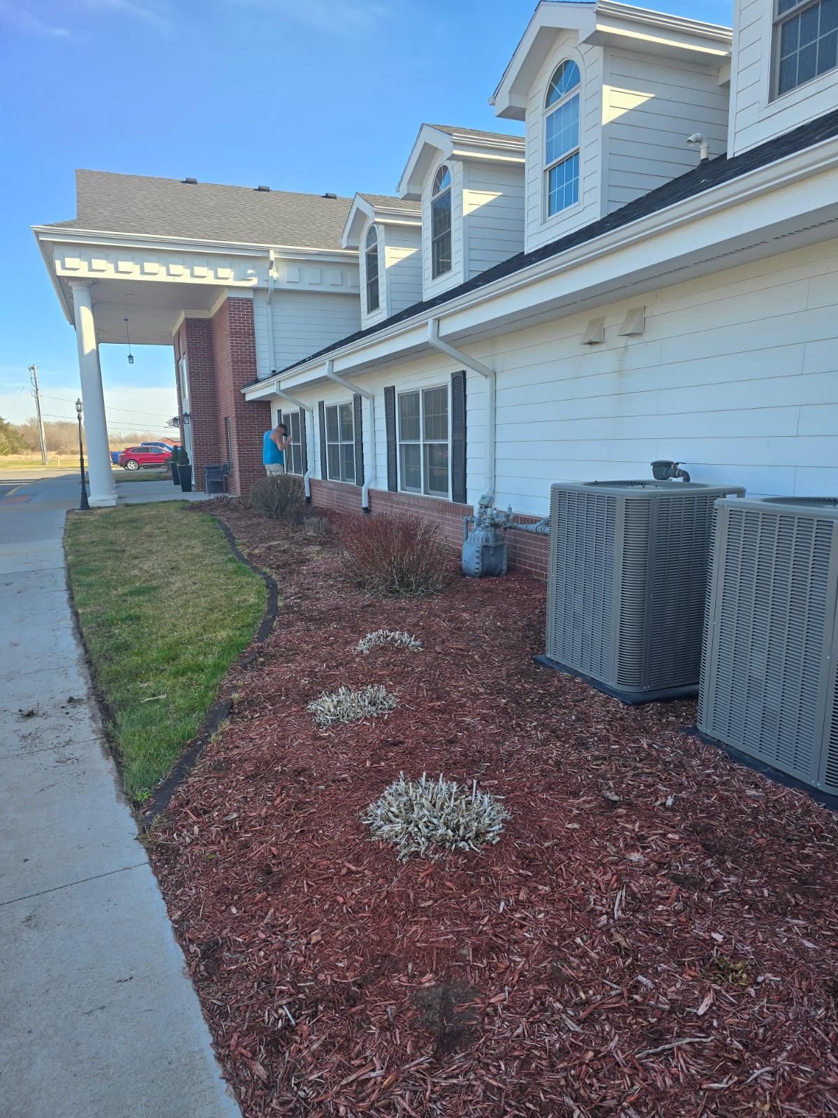 A low-angle view of a white, multi-story building with a red brick base, mulch landscaping, and two large HVAC units.