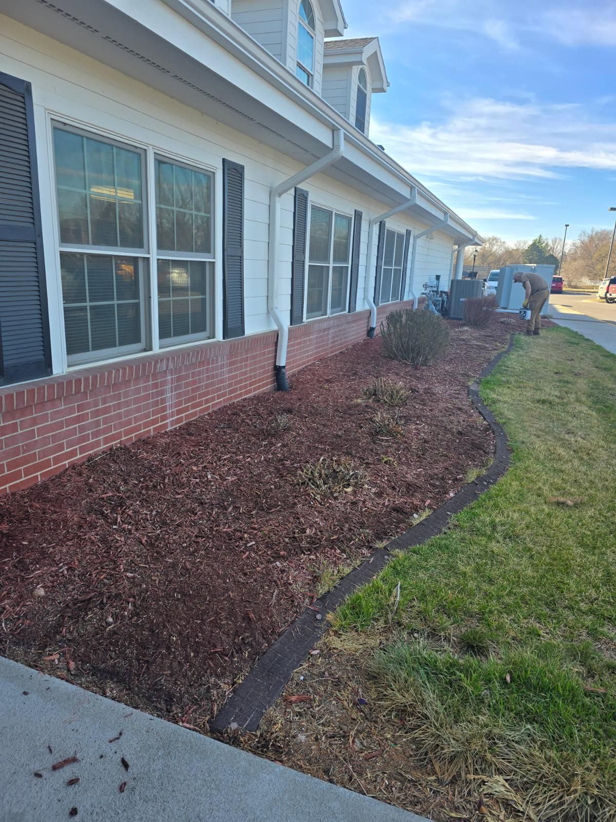 A house with white siding and a brick base, featuring brown mulch in the front yard and a person working in the distance.