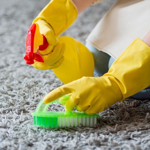 A person wearing yellow gloves is cleaning a carpet with a brush and spray bottle.