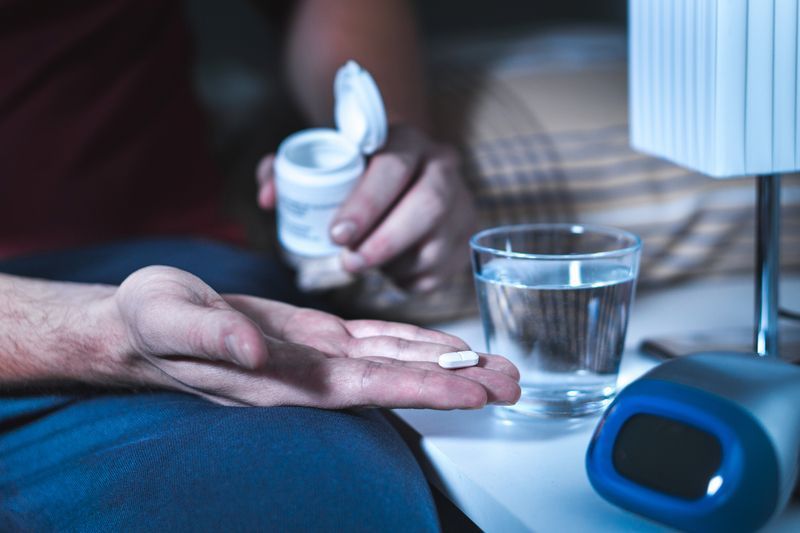 Person taking a pill with a glass of water and alarm clock on a nightstand.