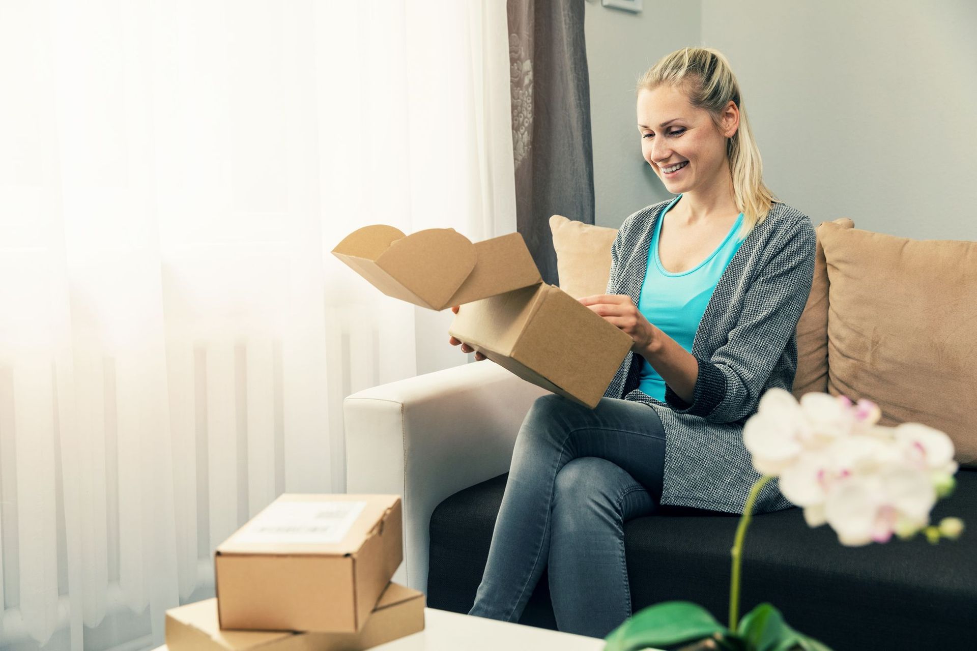 Woman opening a cardboard box on a couch, smiling. Boxes on table, orchid in foreground, window in background.