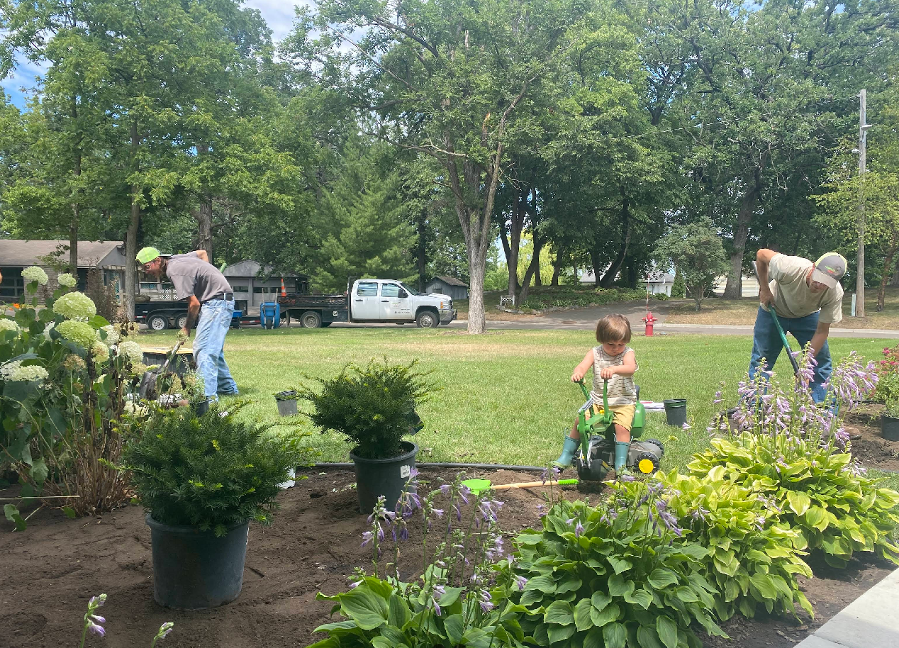 People gardening in a sunny yard, with a child using a toy lawnmower.