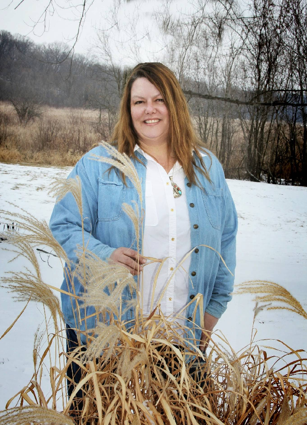 Woman in denim jacket and white shirt smiles in snowy field, holding tall grass.