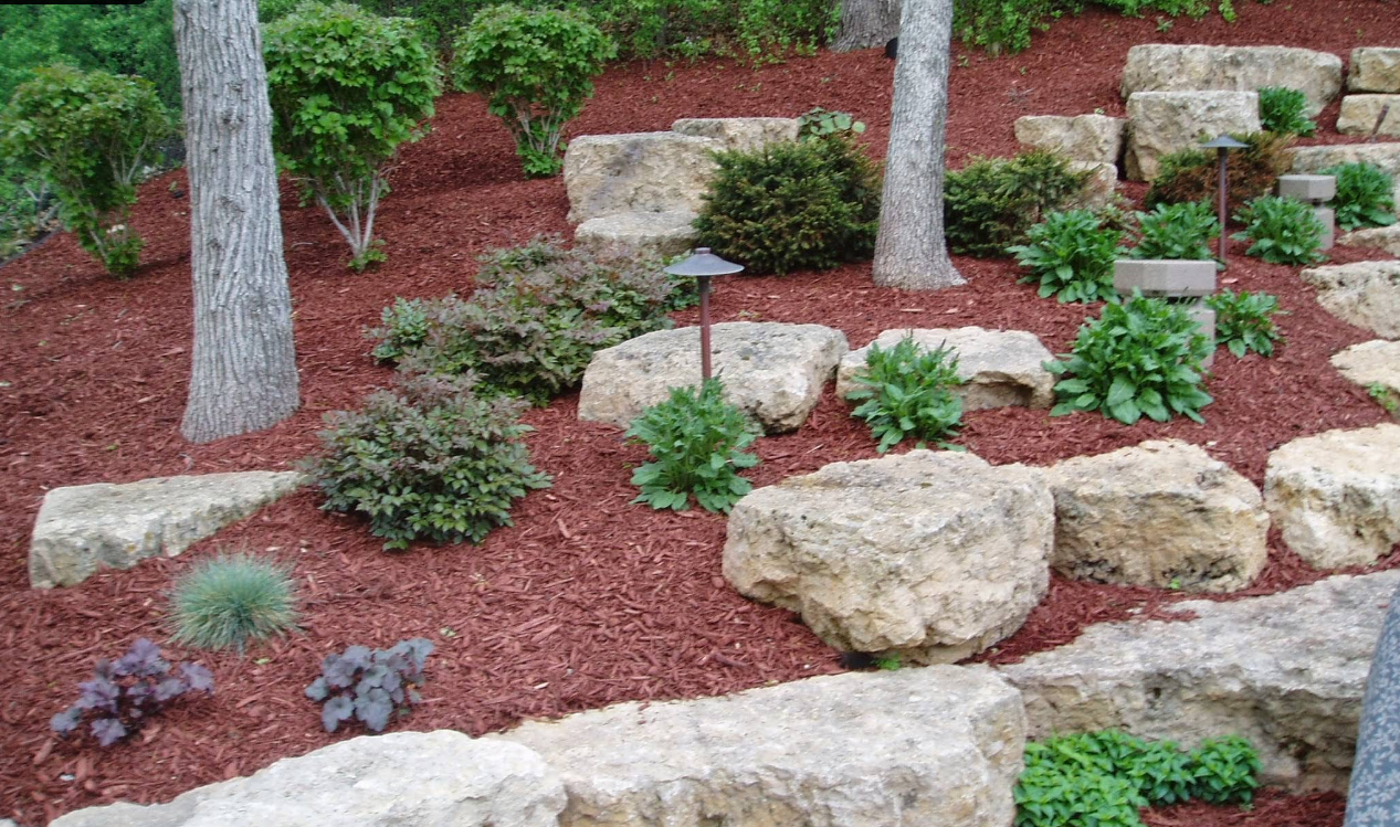 Landscaped hillside with red mulch, large stones, trees, and various green plants.