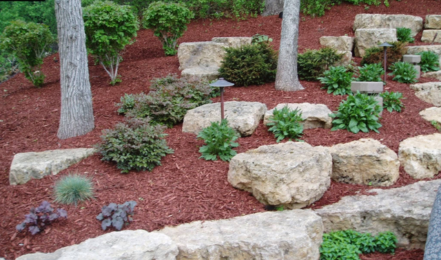 Red mulch and stone retaining wall with shrubs and small trees.