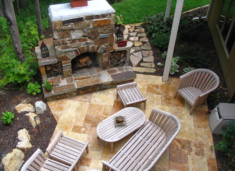 Patio with stone fireplace, wood furniture, and stone paving. Lush greenery surrounds the space.