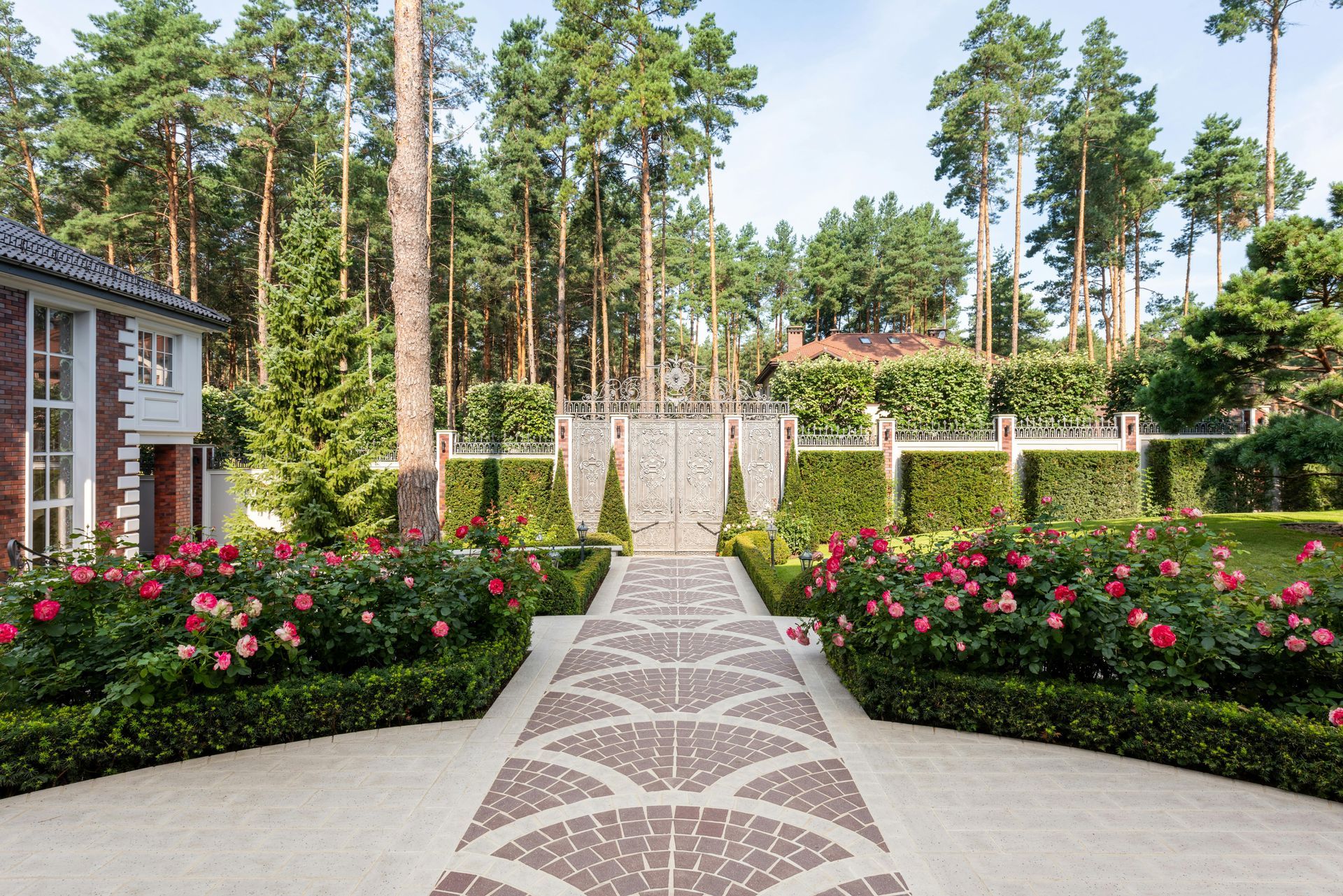 A landscaped garden path leading to ornate iron gates, framed by rose bushes and manicured hedges in a wooded setting.