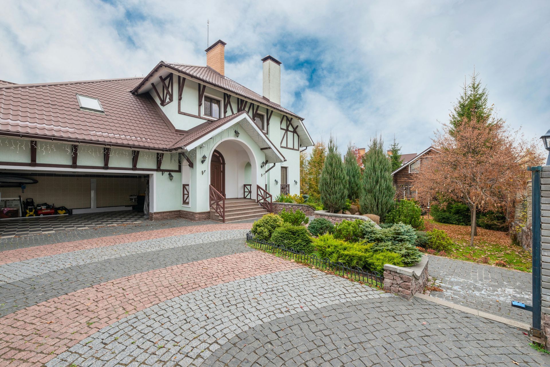 Large house with a cobblestone driveway, arched entryway, and landscaped garden under a cloudy sky