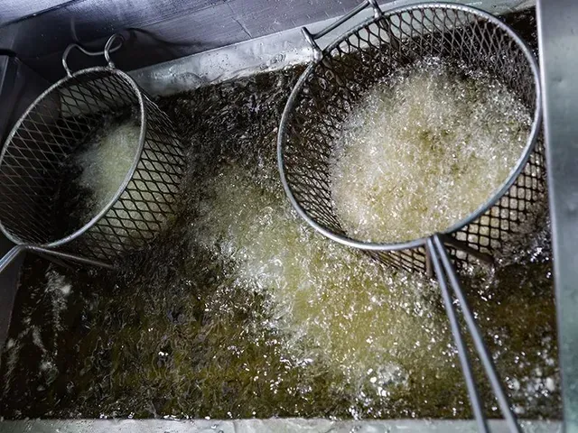 Due cestelli metallici per friggere immersi in olio bollente color marrone dorato all'interno di una friggitrice professionale.