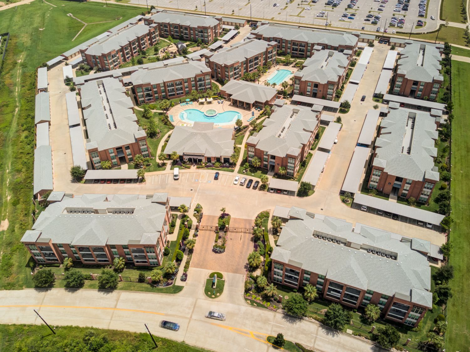 Aerial view of an apartment complex with multiple buildings, a pool, and parking, surrounded by green space.