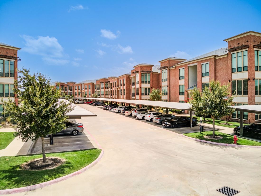 Apartment complex with covered parking. Brick buildings, cars parked under canopies, and green grass.