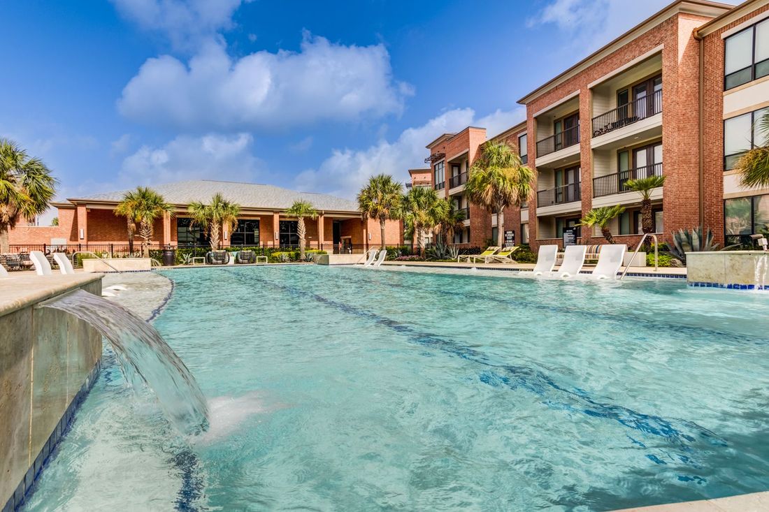 Swimming pool with waterfall feature and brick apartment buildings under a blue sky.