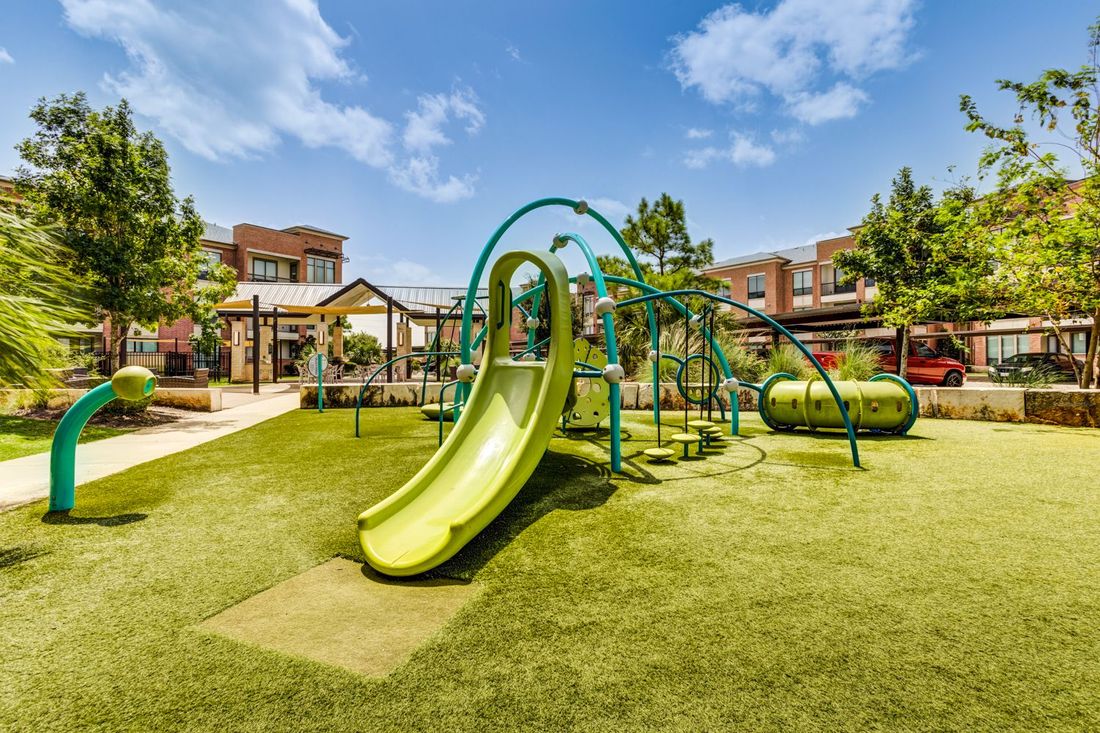 Playground with green slide and structures on artificial turf in a sunny outdoor setting.