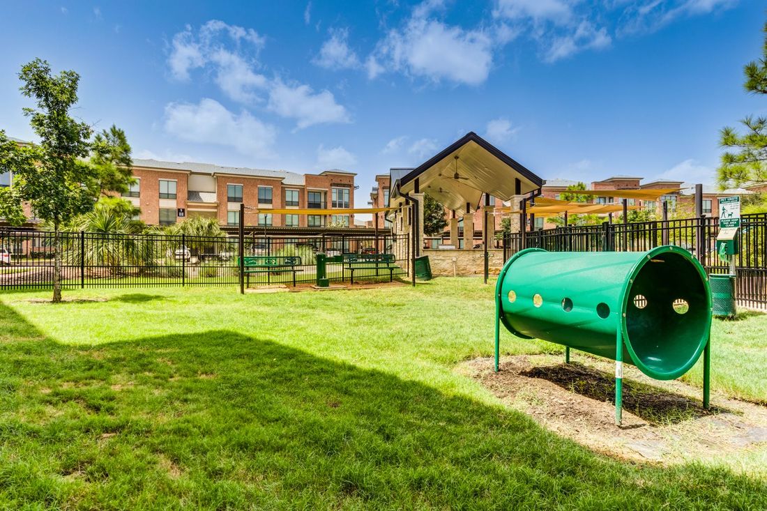 Dog park with green tunnel, grass, fence, and multi-story apartment buildings under a blue sky.