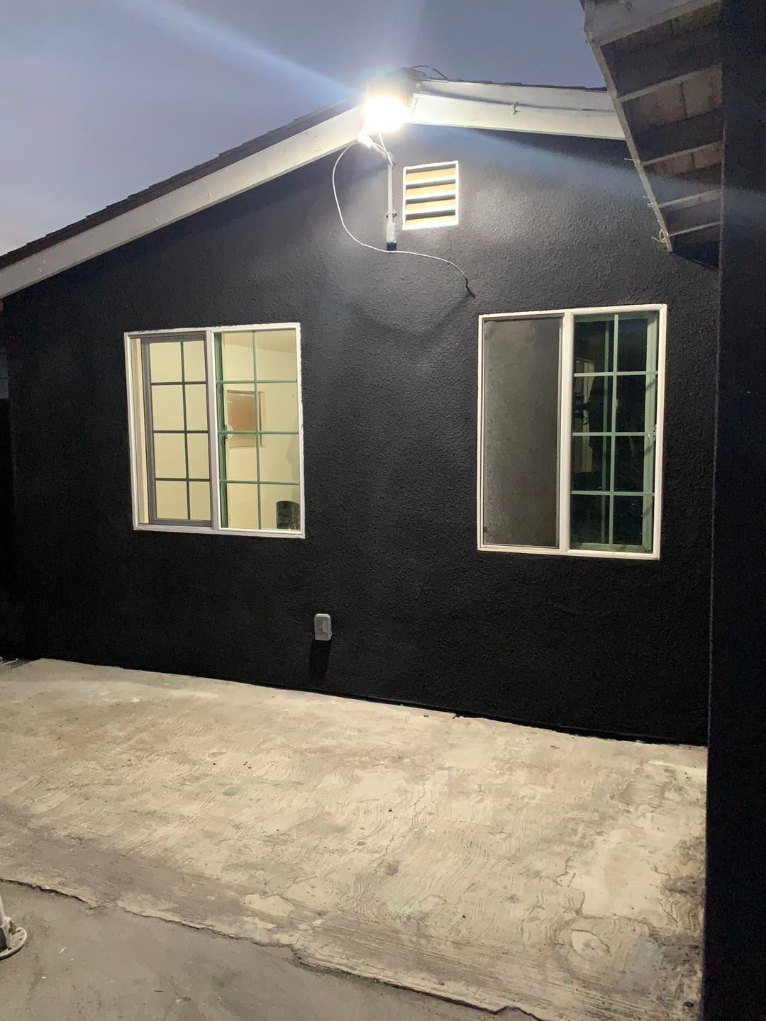 Small black exterior wall with two windows, a light fixture, and a concrete patio at dusk