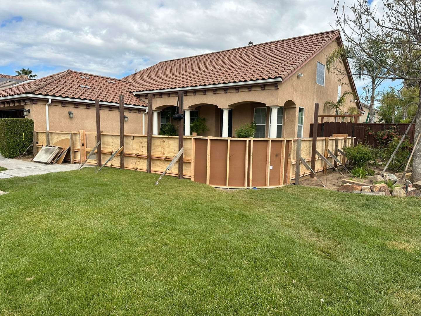 Backyard with a single-story house, tiled roof, fenced patio, and green lawn under cloudy skies