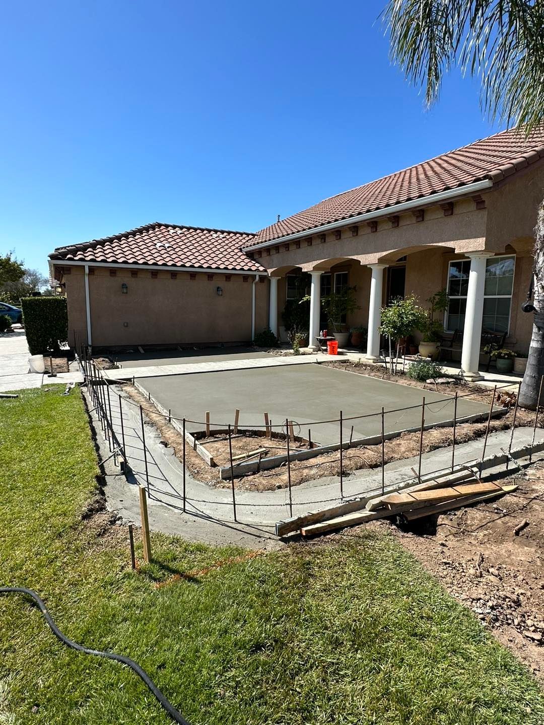 Backyard concrete patio under construction beside a stucco house and garage, with rebar and forms visible.