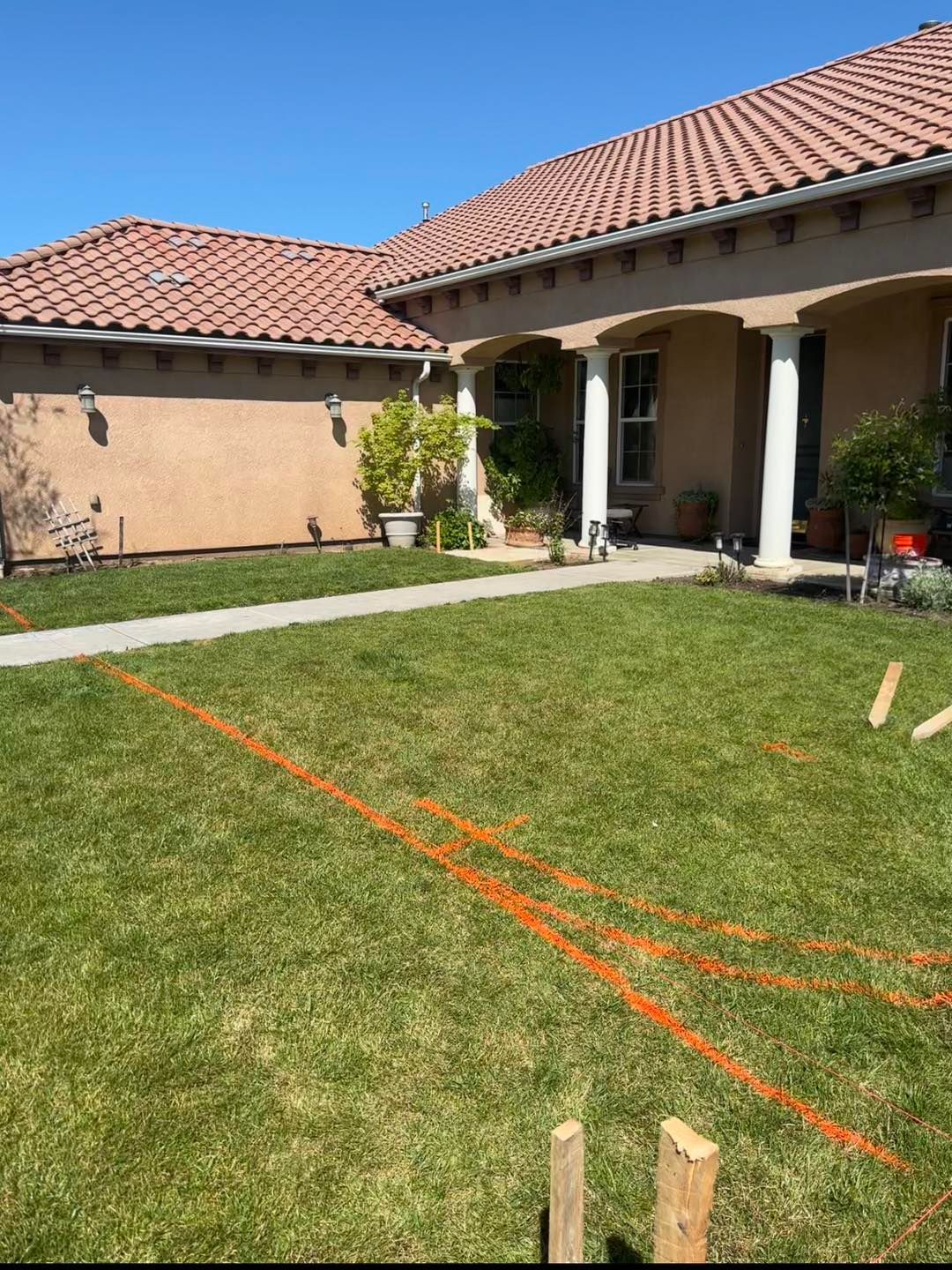 Backyard lawn with orange string lines, patio, and a house with a red tile roof under a clear blue sky