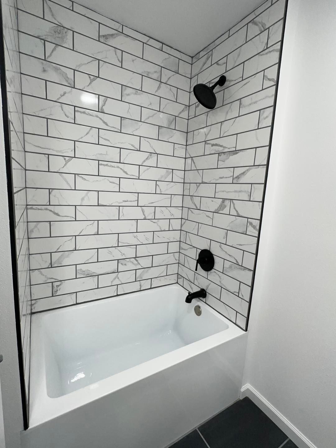 Bathtub alcove with white subway tile walls, black fixtures, and gray walls and floor