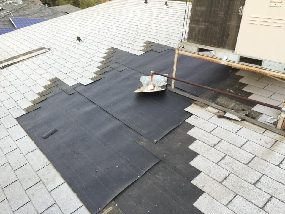 Roof under repair with black underlayment patches on light gray shingles and a small tray of tools near the ridge